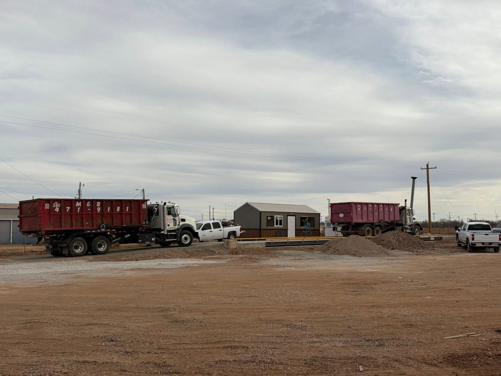 Two dump trucks parked near a small building on a dirt lot under an overcast sky.