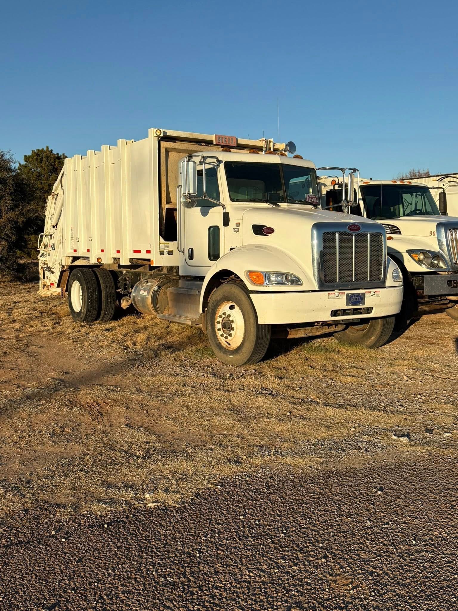 White garbage truck parked on dry, brown ground under a blue sky.