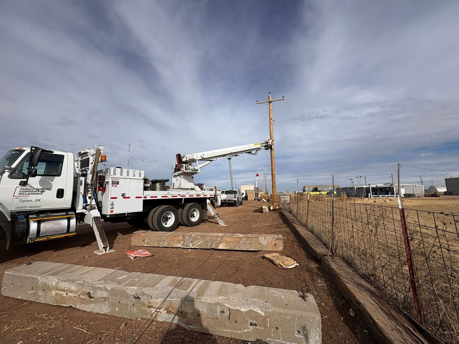 Utility truck with extended boom working on a wooden power pole. Cloudy sky, brown field, and concrete blocks.