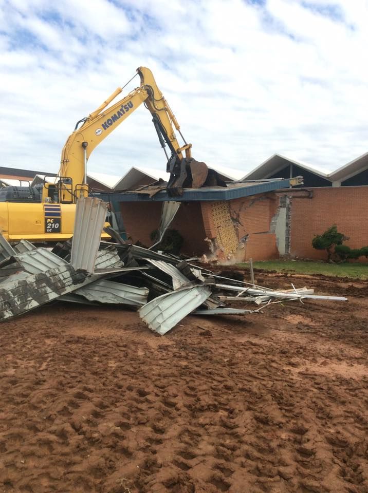 Yellow excavator demolishing a brick building with a corrugated metal roof on a muddy construction site.