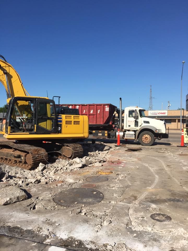 Construction site with a yellow excavator, red dumpster, and white truck on a broken concrete surface under a blue sky.