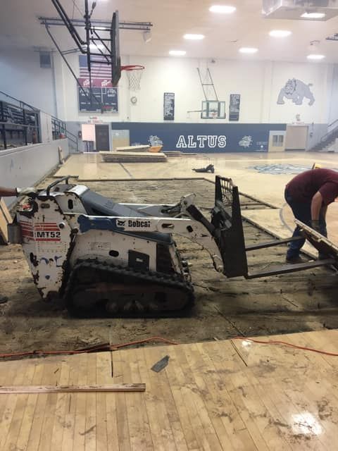 Bobcat skid steer removing a section of a gymnasium floor in Altus, Oklahoma. A person works nearby.