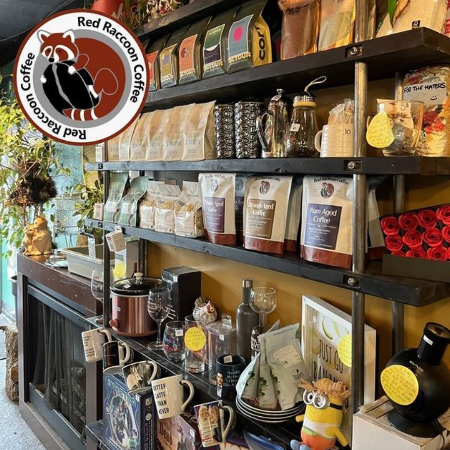 Shelves stocked with coffee bags, mugs, and decor inside a coffee shop. Red raccoon logo in the corner.