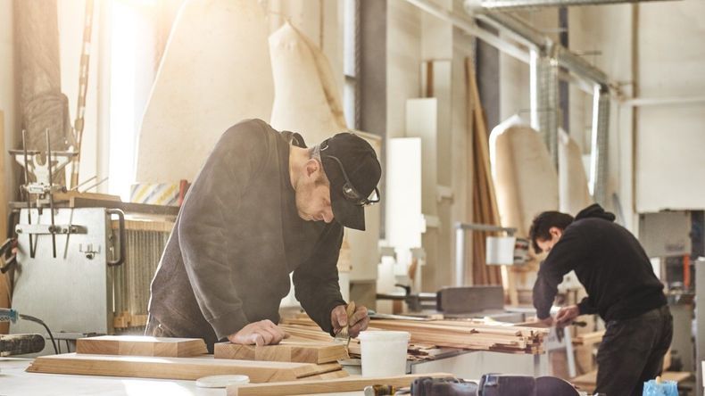 Two workers sanding wooden boards in a bright workshop, with tools and lumber scattered on the table.
