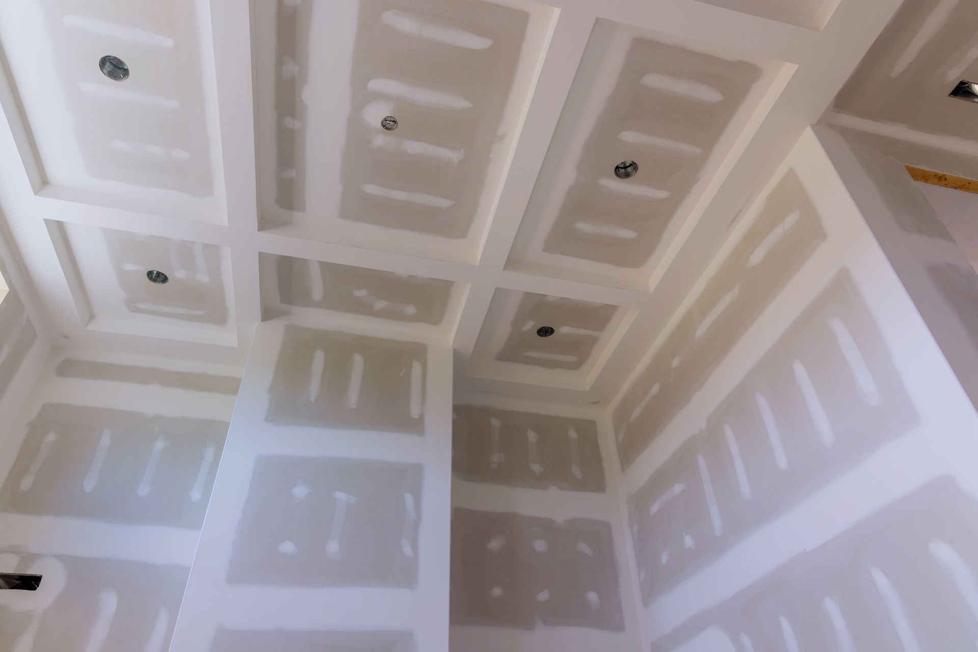 Unfinished drywall ceiling and walls in a room, with taped seams and recessed light openings