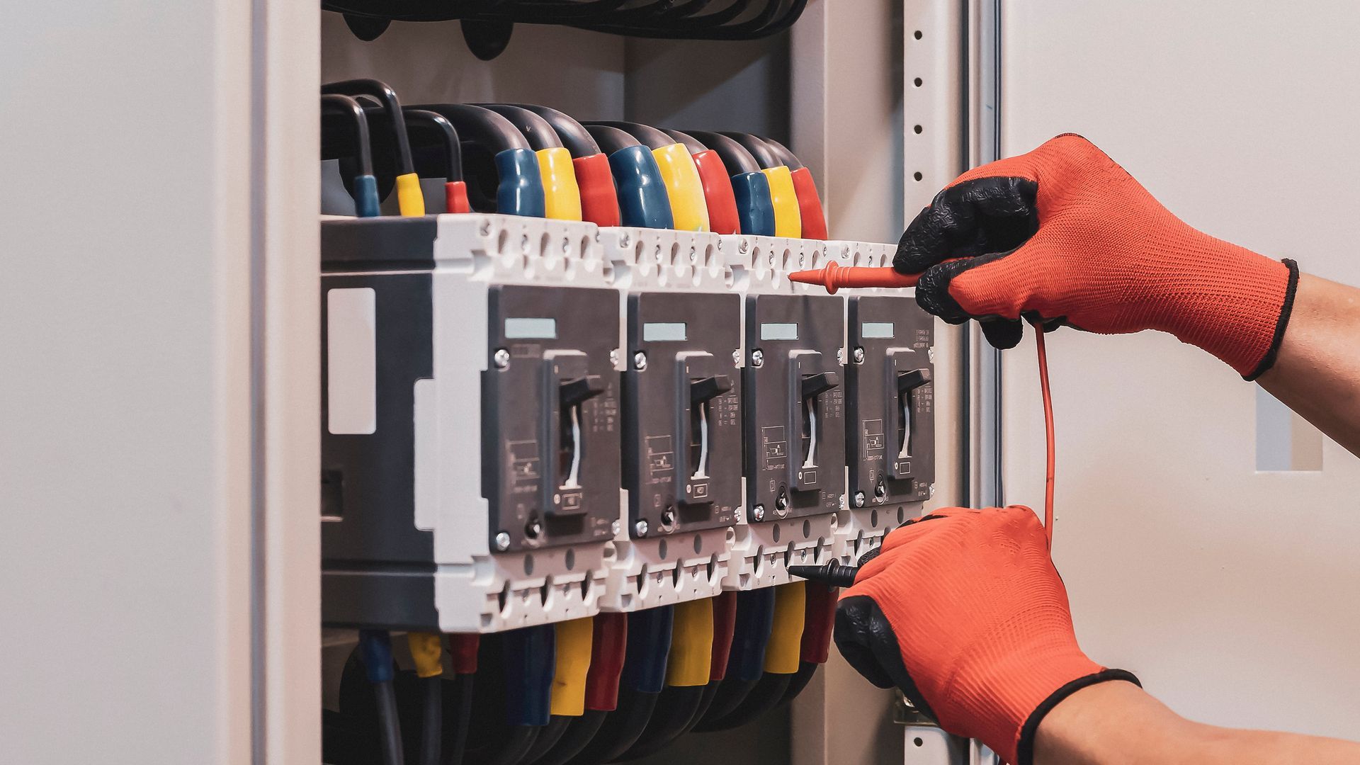 Gloved hands adjusting circuit breakers in an electrical panel, with multicolored wires visible behind.