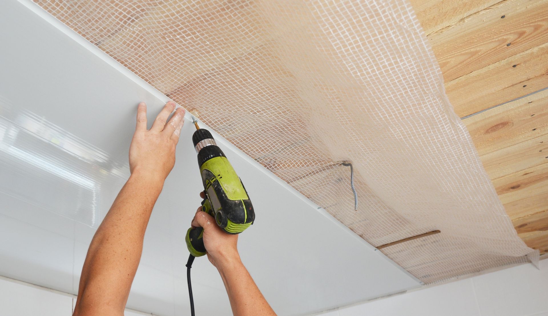 Hands using a green drill to install white ceiling panels in a room.