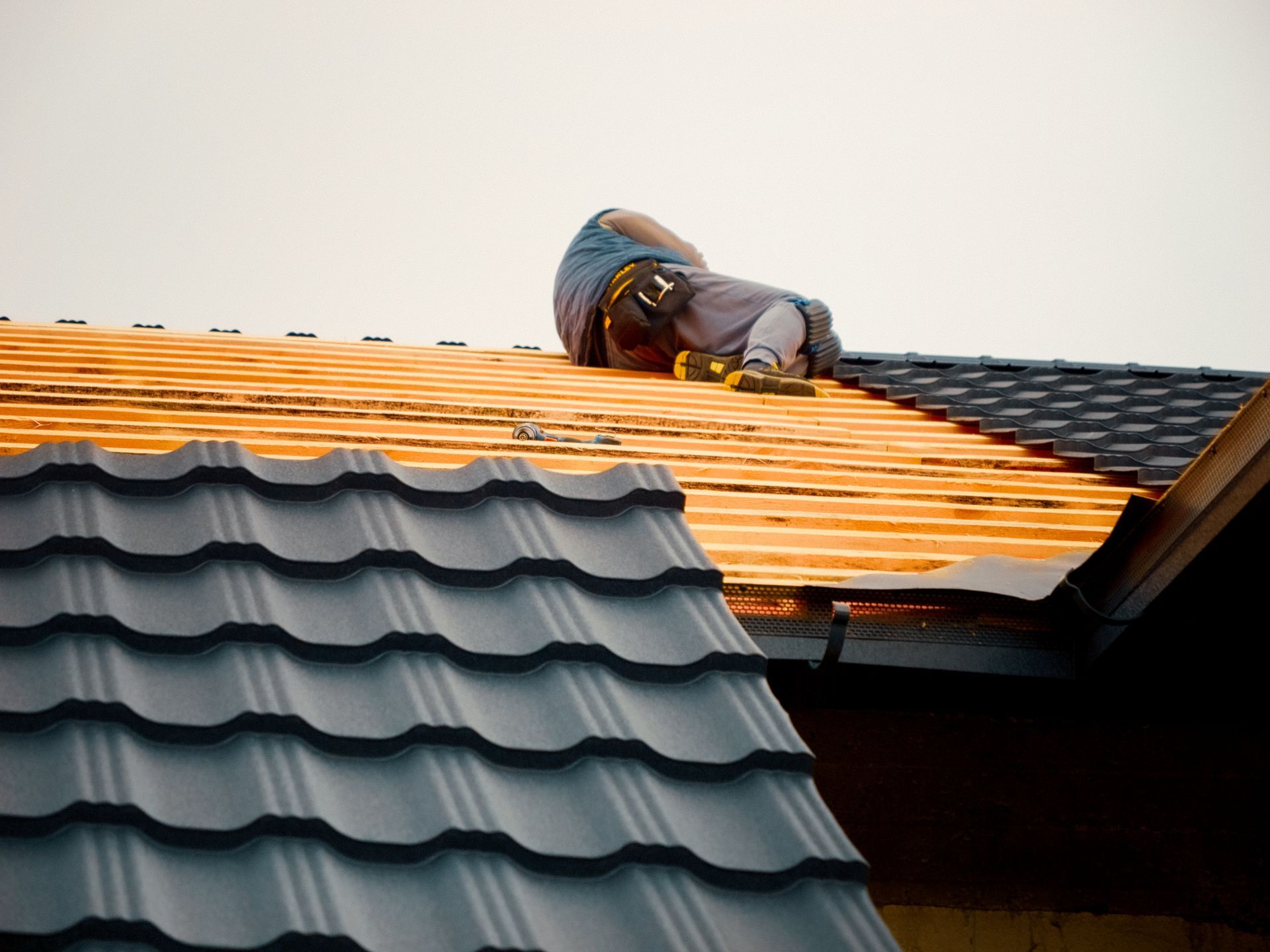Worker kneeling on a roof, installing orange roofing underlayment beside dark gray shingles.