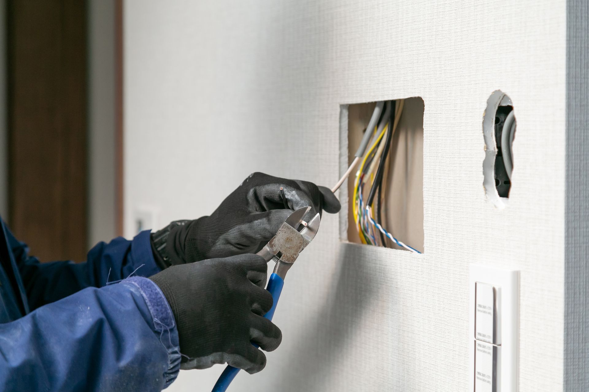 Gloved hands wiring an electrical box in a wall, with exposed cables and tools
