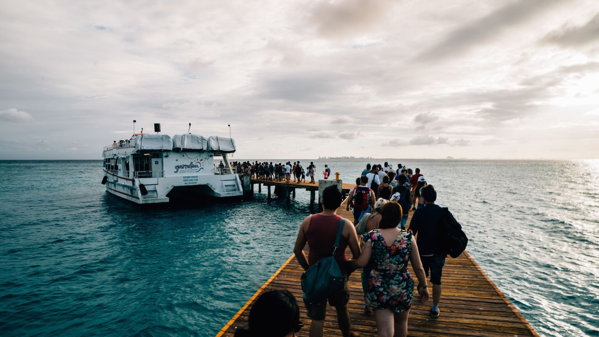 Passengers walking along a wooden pier toward a waiting ferry under a cloudy sky.