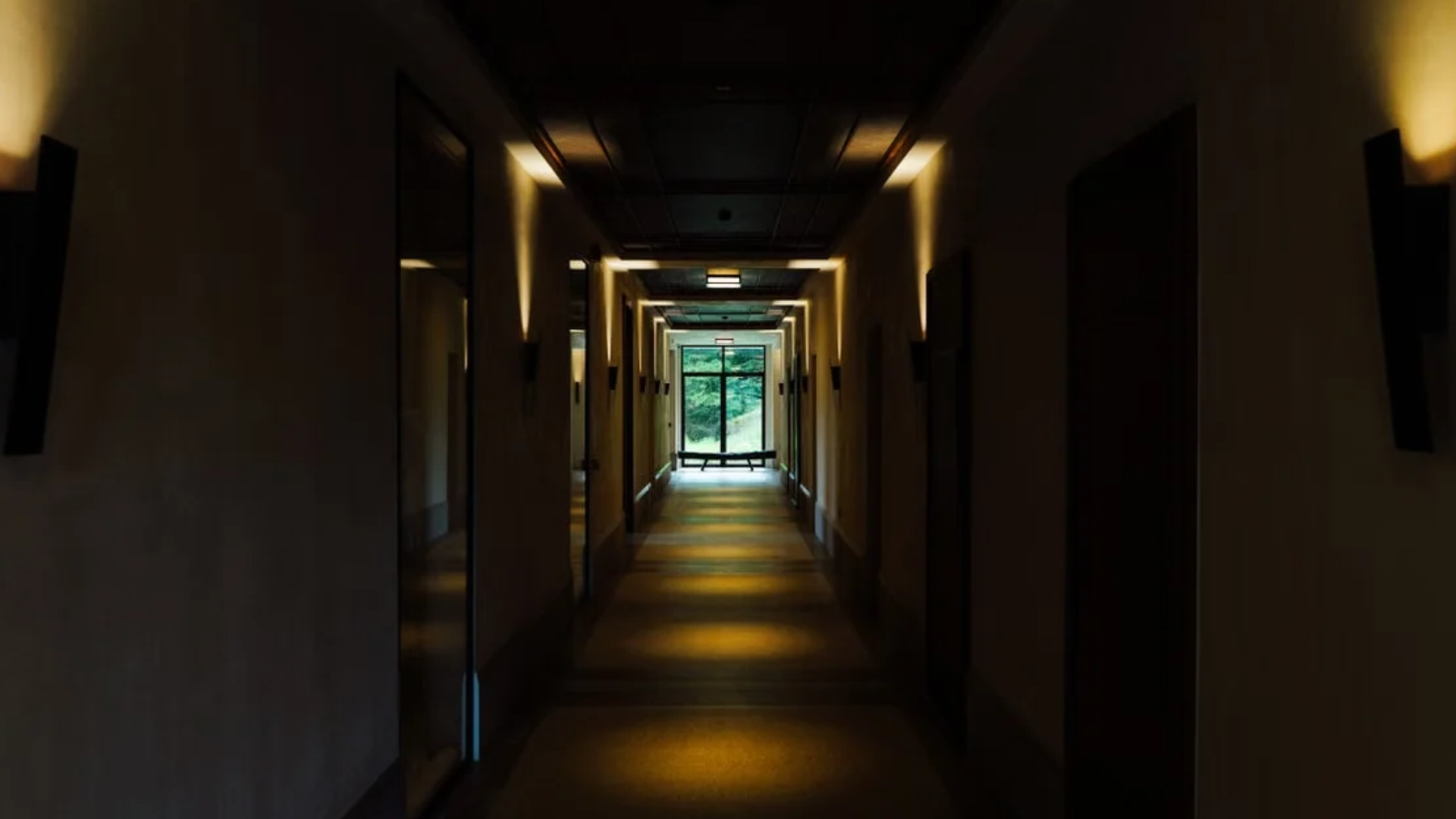 A dim, empty hotel corridor at night, lit by wall sconces, leading to a window at the far end.