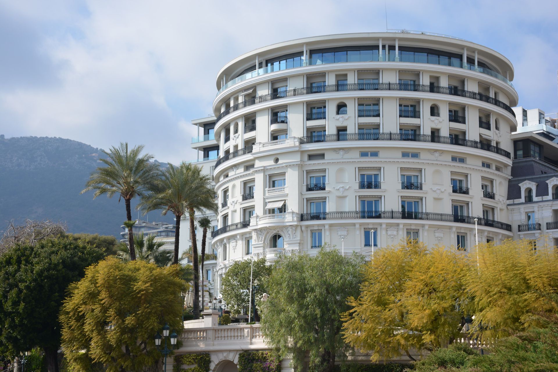 Large white circular hotel building with balconies, framed by palm trees and mountains under a cloudy sky