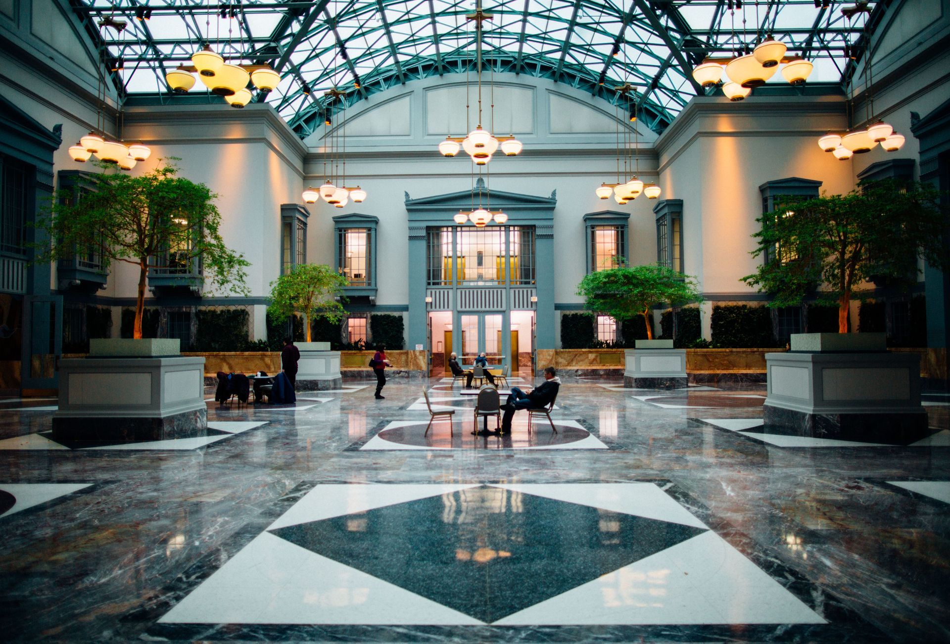 Grand lobby with glass dome, marble floor, potted trees, and a few people seated in the center.