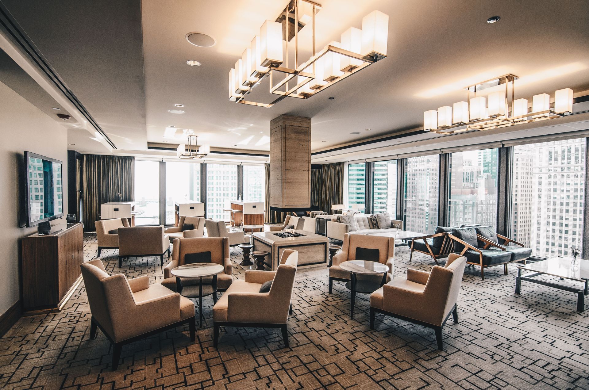 Modern hotel lounge with beige chairs, patterned carpet, large windows, and chandeliers