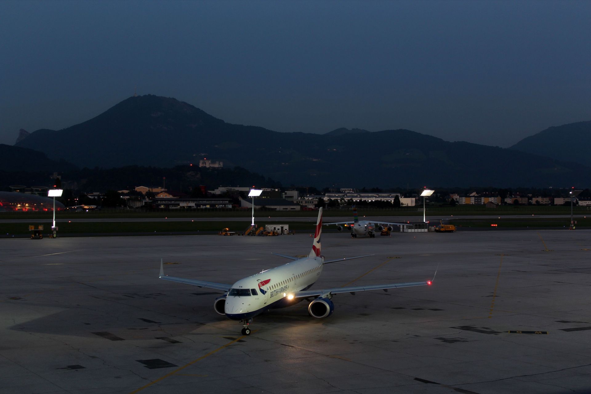 Airplane parked on a dimly lit airport tarmac at dusk, with mountains in the background.