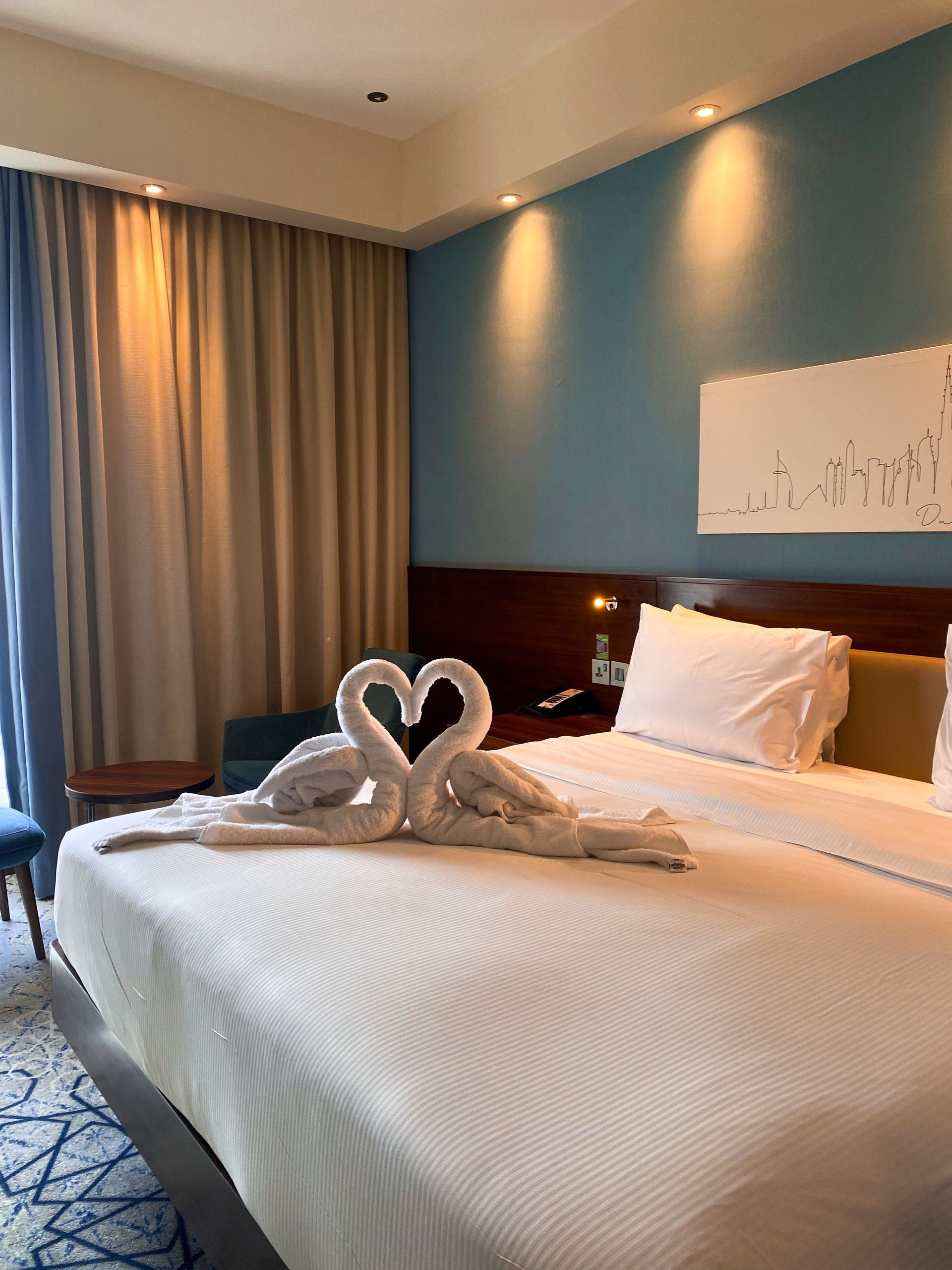 Hotel room with a neatly made bed, heart-shaped towel arrangement, blue wall, and curtains near a window.