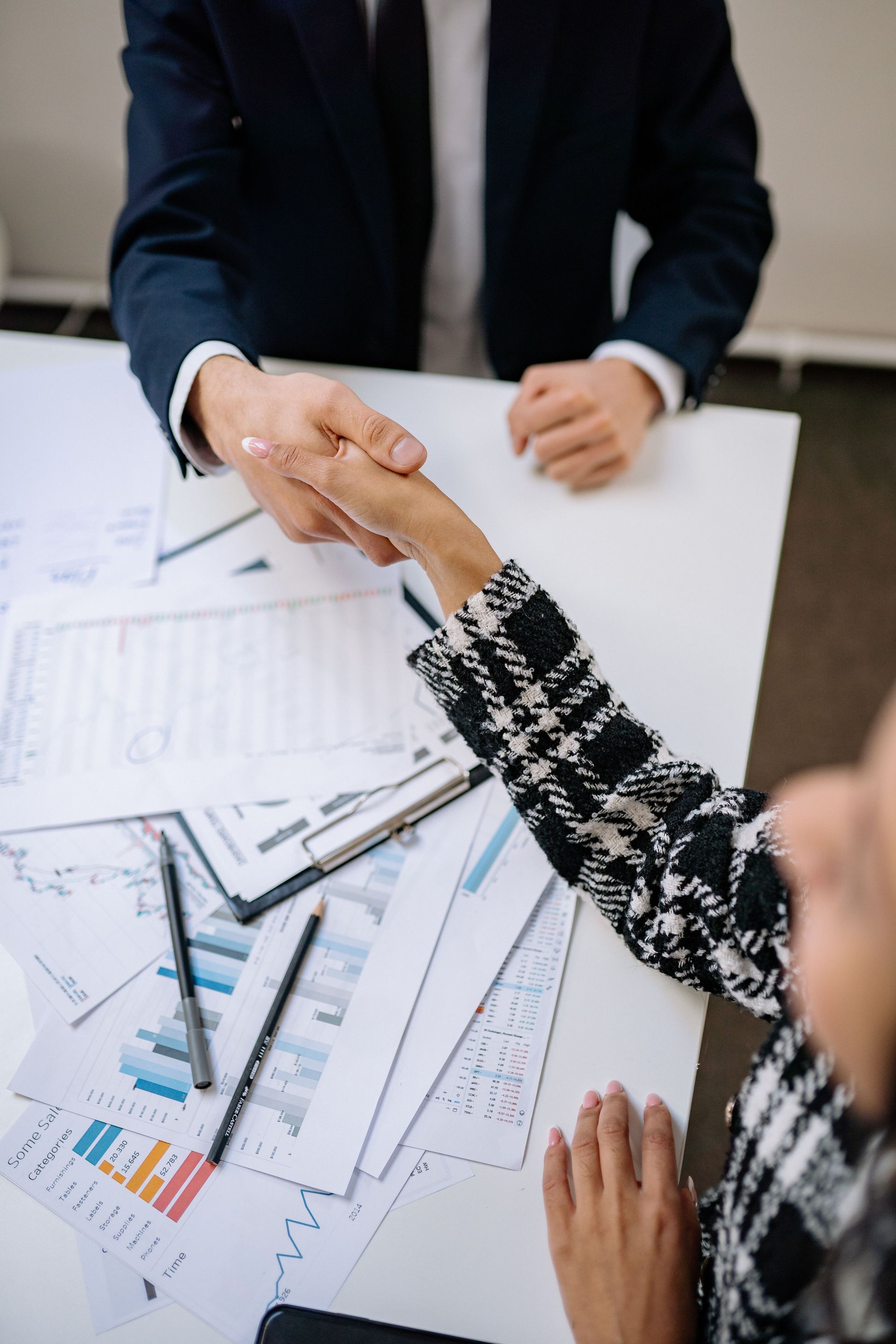 Two people shake hands across a desk with a laptop and documents in a bright office