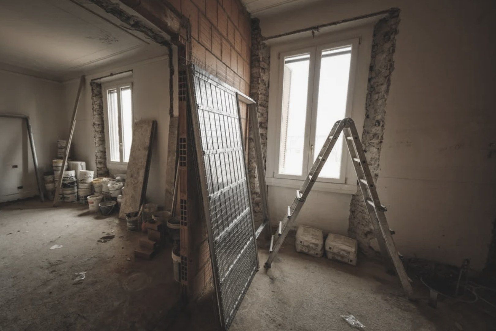 A room stripped back for repair works, with bare masonry around the windows, a stepladder and building materials on a concrete floor.