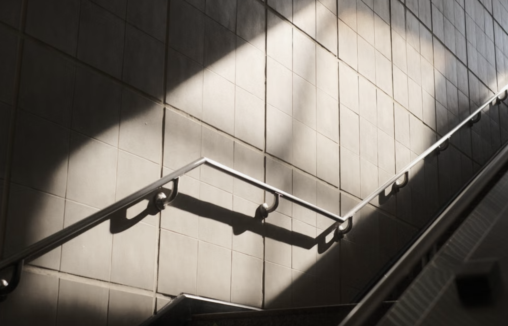 Daylight falls across a tiled stairwell wall, casting the shadow of a steel handrail.