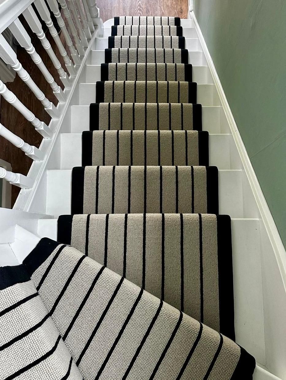 Staircase with striped carpet runner in neutral tones and black borders. White risers and banister.