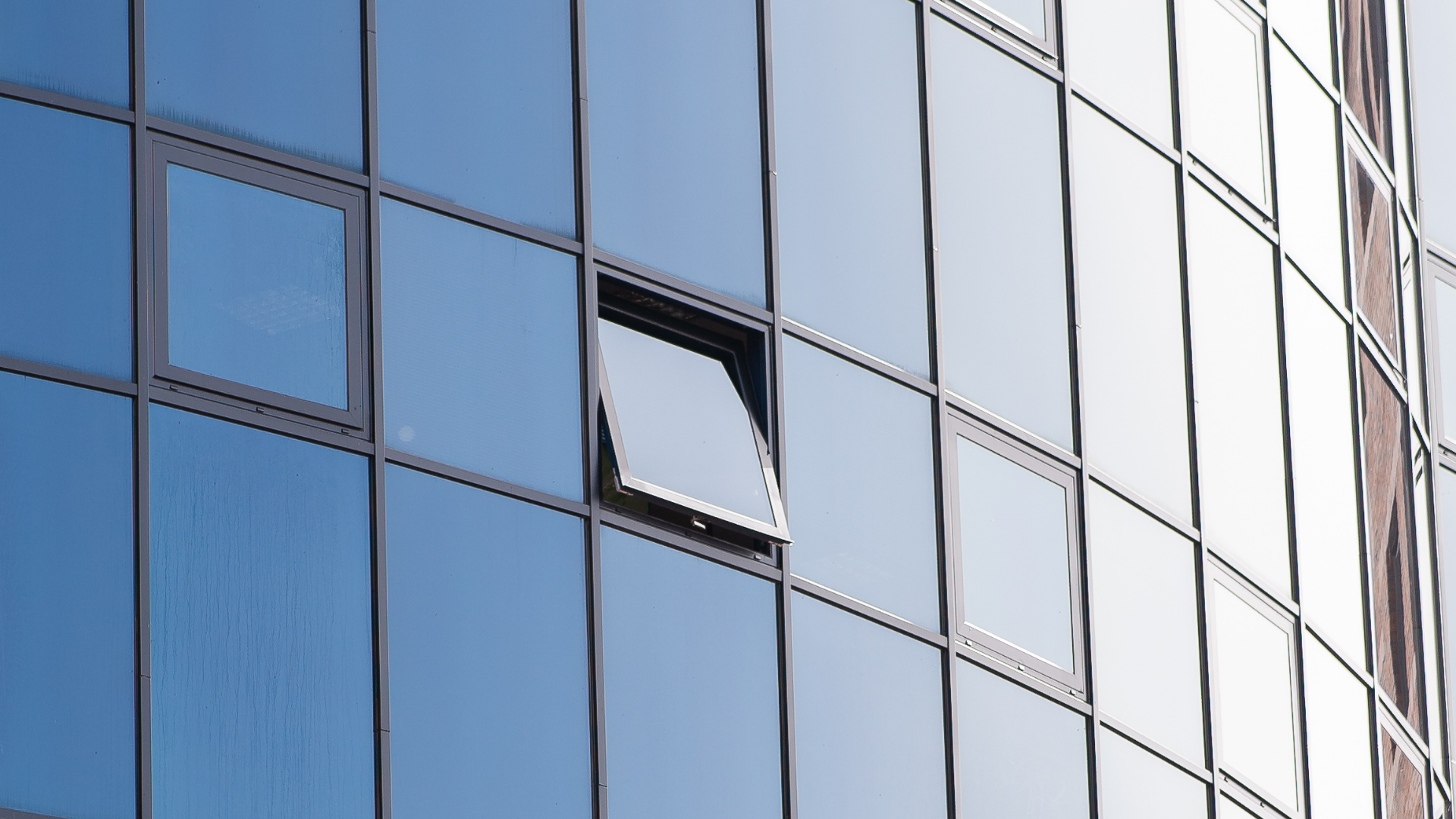 A modern glass-paneled building facade with a single window open among many closed panes.