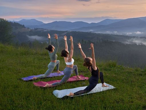 Three women are practicing yoga on top of a grassy hill.