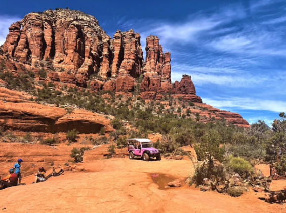 A pink jeep is parked on a dirt road in front of a large rock formation.