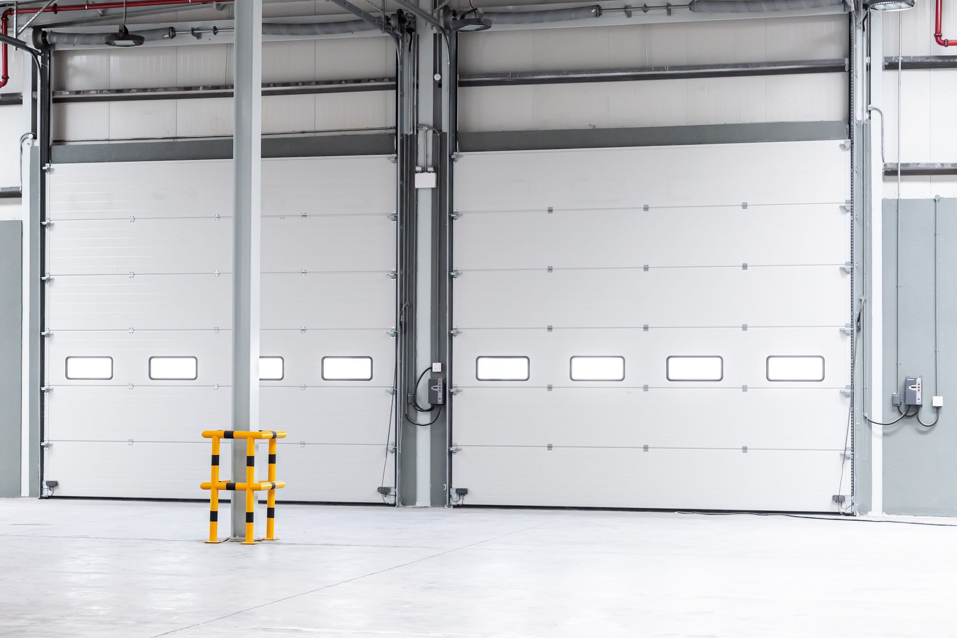 Two closed white industrial doors in a warehouse, with a yellow and black safety barrier at the center.