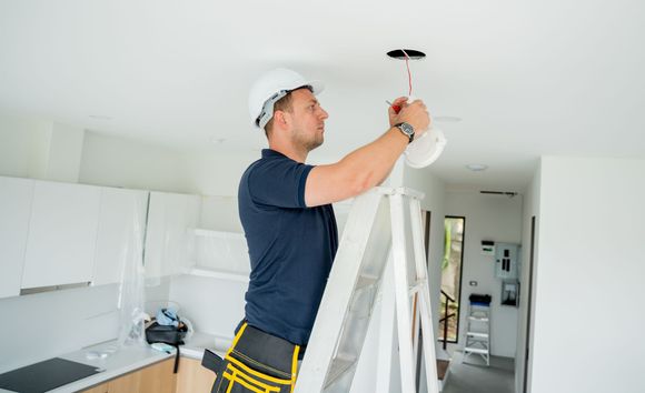 Electrician on a ladder installing a ceiling light fixture in a white room.