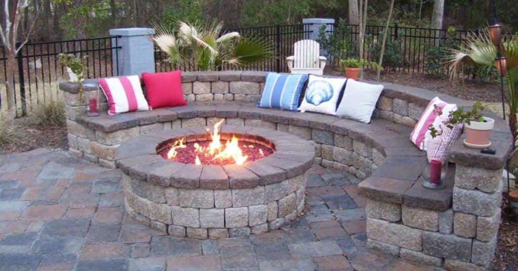 A stone fire pit surrounded by a curved stone seating wall with colorful throw pillows on a paved patio.