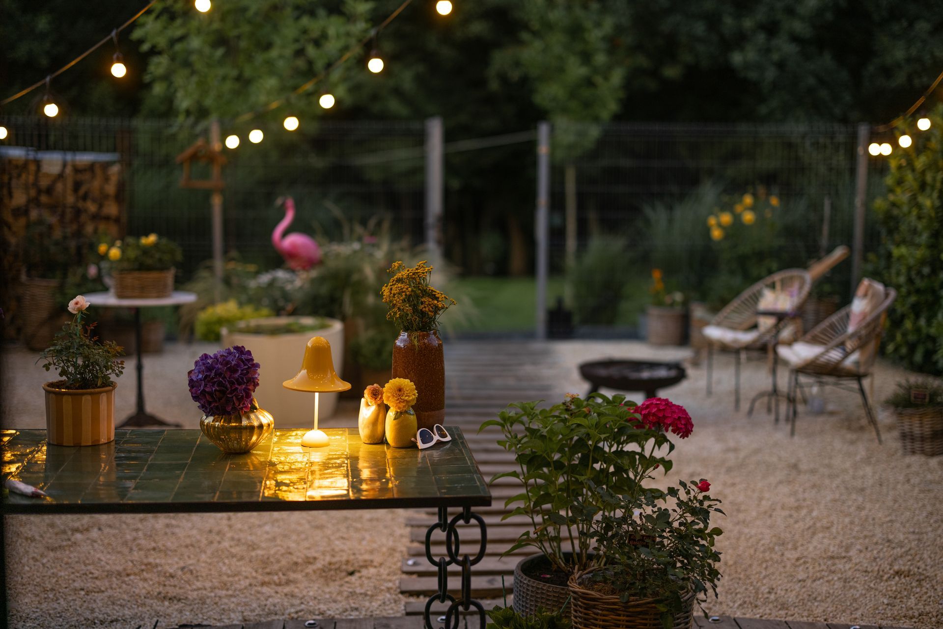 Cozy outdoor patio at twilight, featuring string lights, a table with a glowing lamp, plants, and wicker chairs.