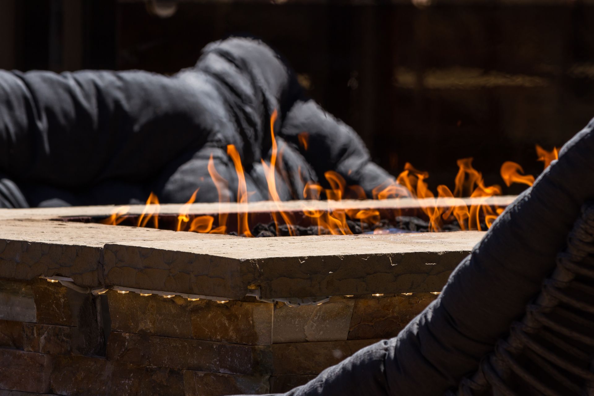Flames rise from a stone fire pit with people in dark jackets sitting around it outdoors.