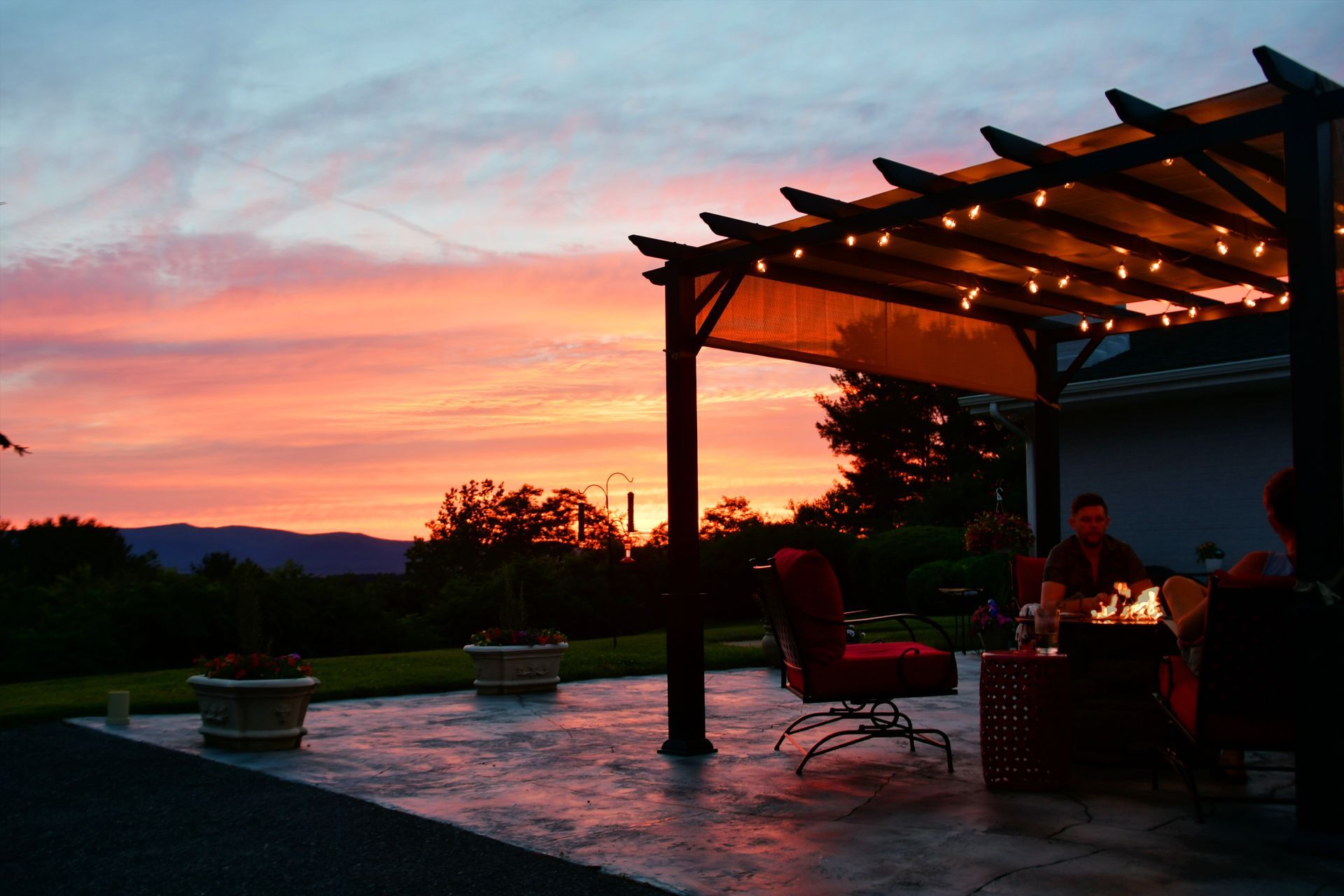 A sunset view from a patio with a pergola, string lights, and people sitting around a fire pit.