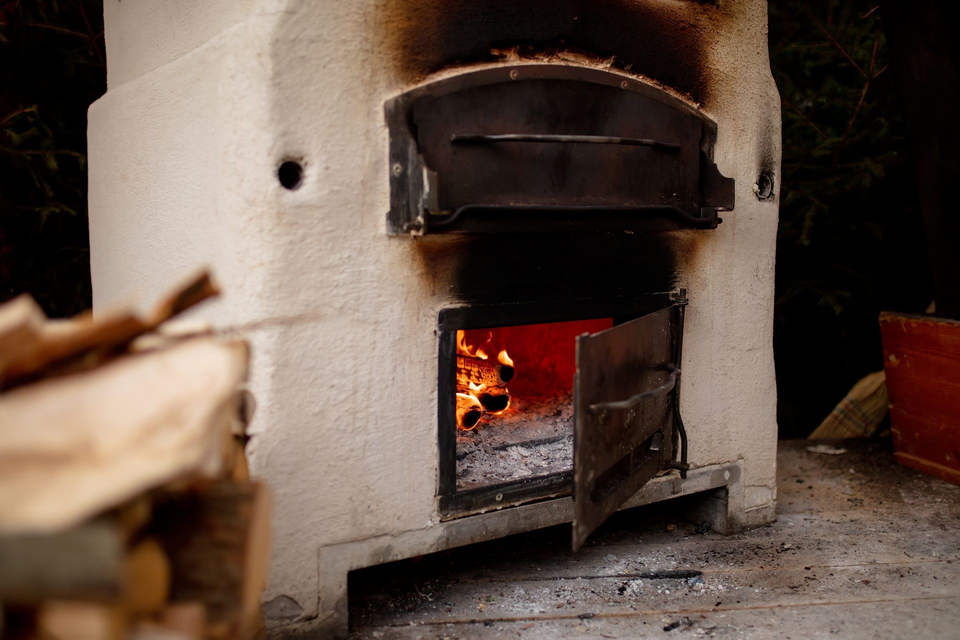 A masonry wood-fired oven with a fire burning in the lower chamber and a stack of firewood in the foreground.