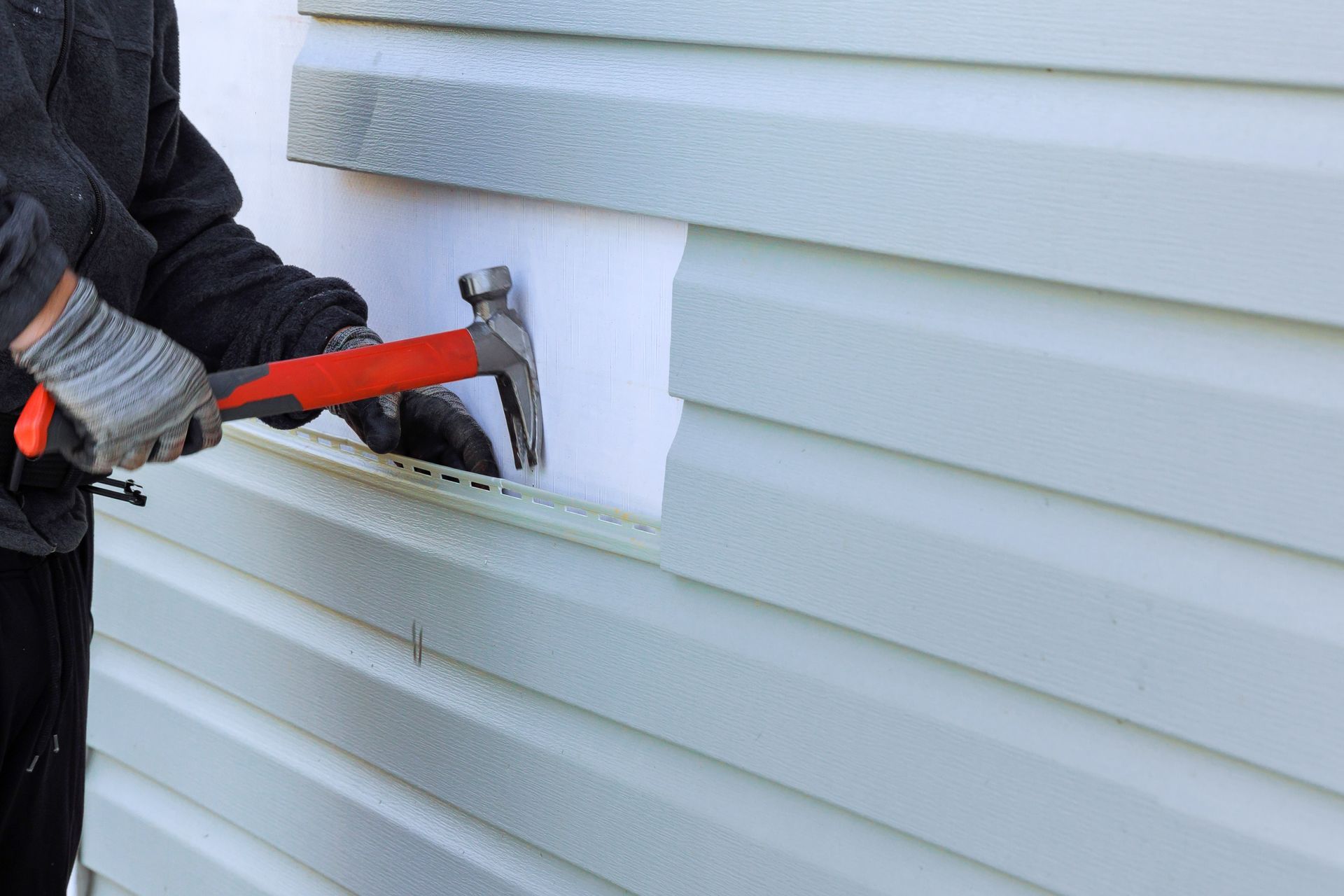 A worker uses a hammer to install light green vinyl siding panels onto a white insulated wall.