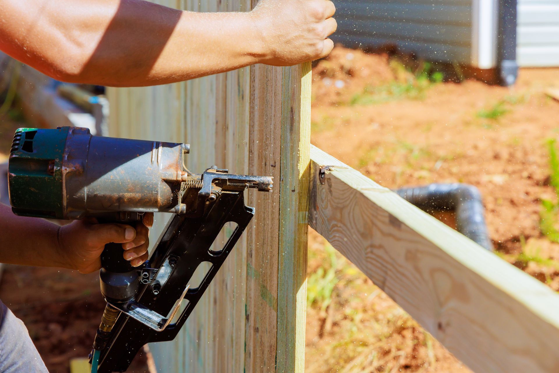A person uses a nail gun to attach a wooden board to a fence rail outdoors.