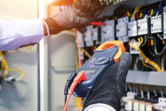 Electrician using a handheld tester on an open electrical control panel with wires and circuit breakers