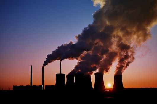Industrial smokestacks releasing thick smoke at sunset, silhouetted against an orange and purple sky