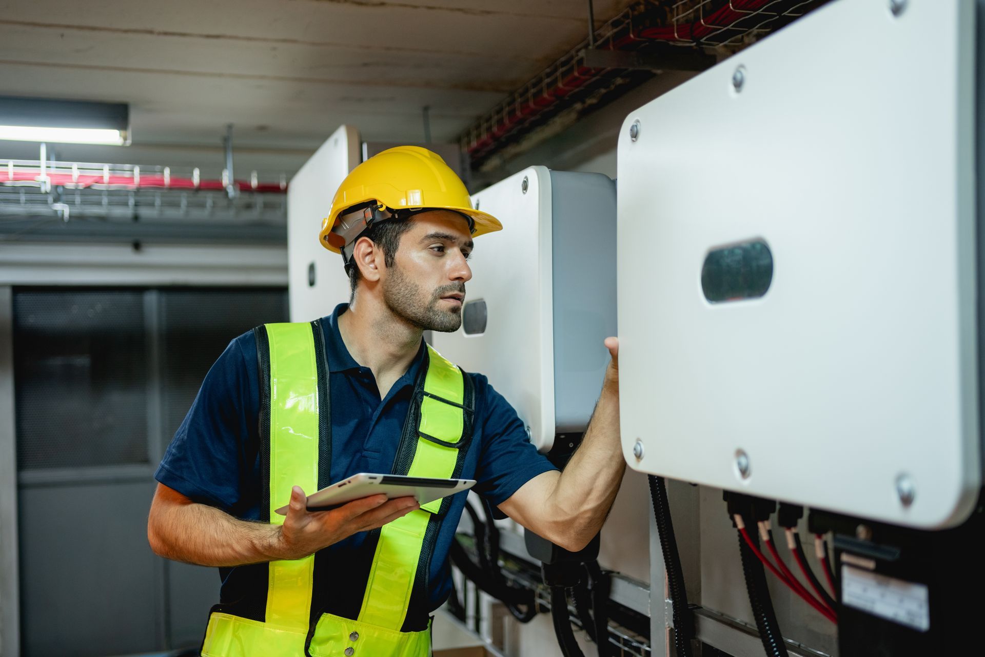 Technician in yellow vest and hard hat inspecting electrical equipment with a tablet.