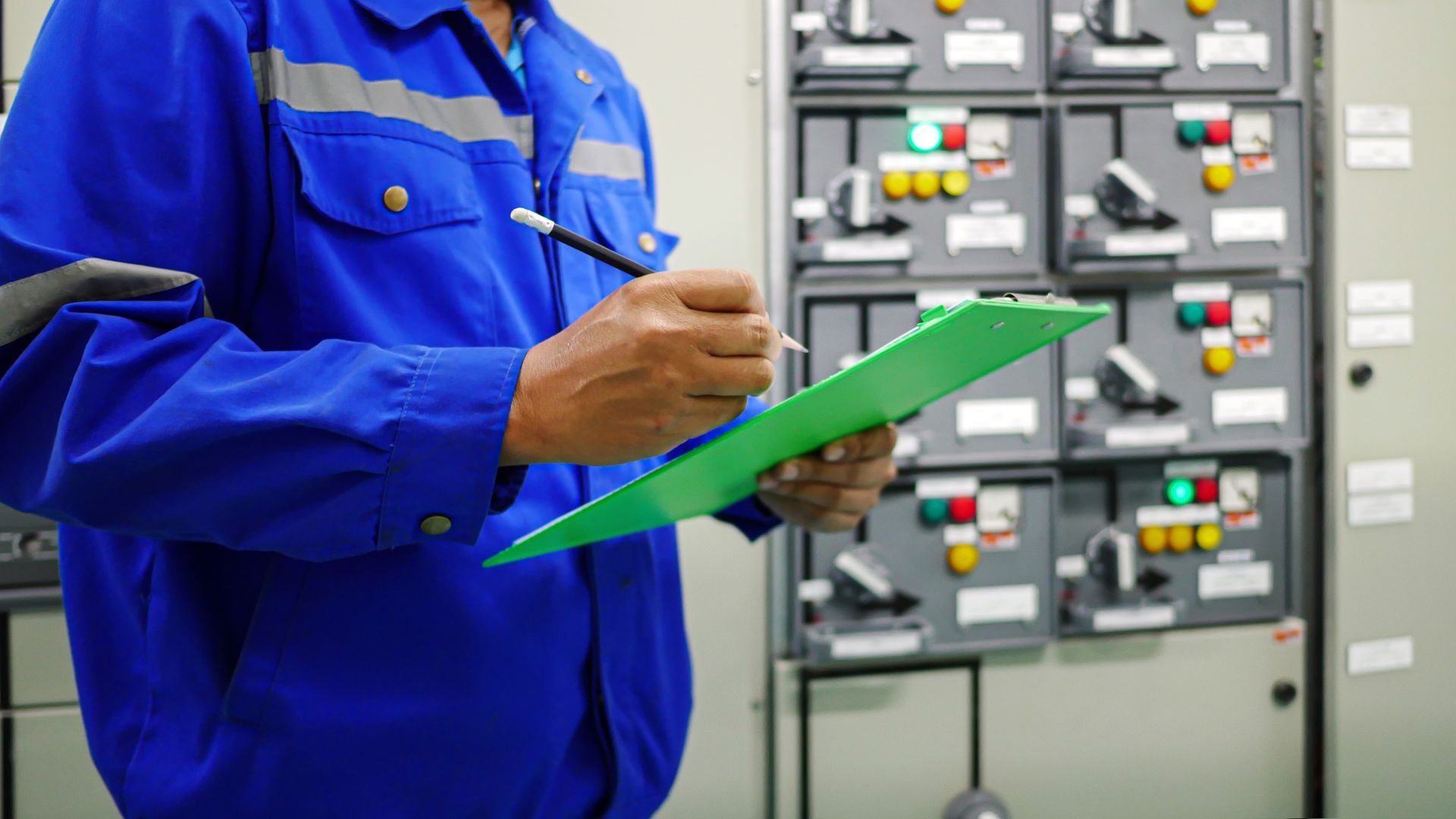 Technician in blue coveralls inspecting electrical control panels with a green clipboard