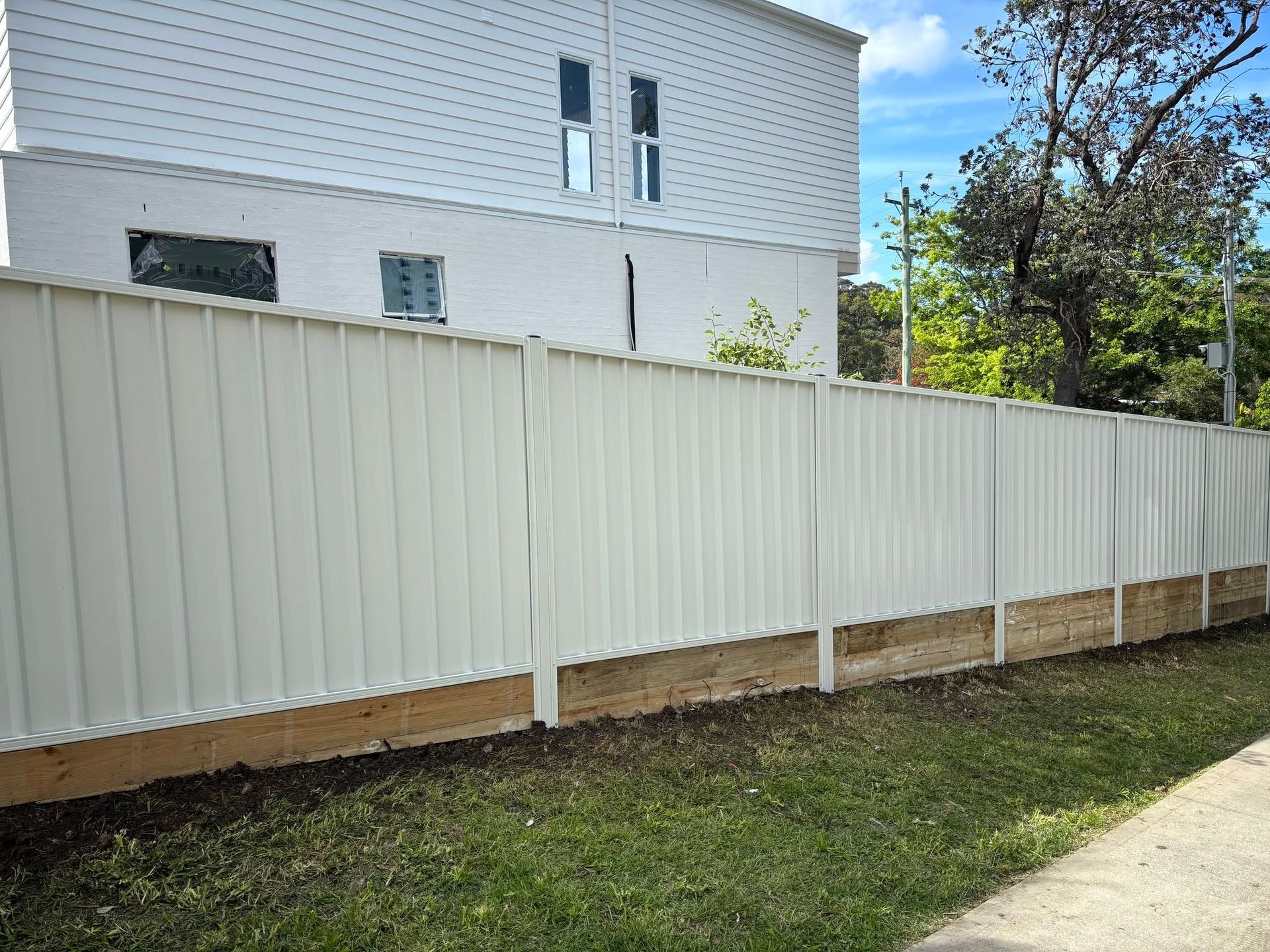 White fence along a grassy yard beside a light-colored building and sidewalk under a clear blue sky