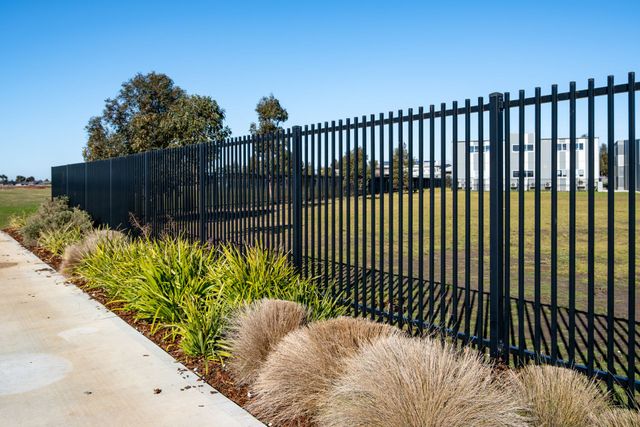 Black metal fence along a sidewalk beside a grassy field and shrubs under a clear blue sky