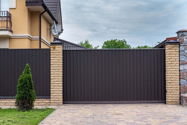 Dark metal gate between beige stone pillars beside a modern house and paved driveway