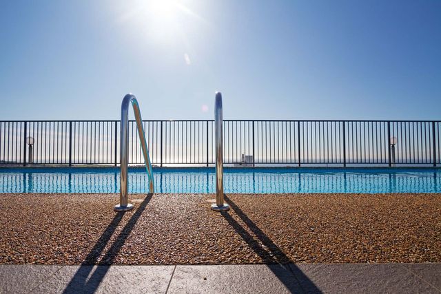 Sunlit seaside promenade with metal railing, pebble ground, and ocean beyond