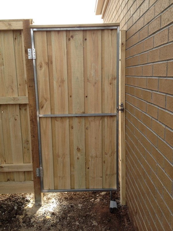 Wooden gate set between a fence and brick wall in a narrow outdoor passage.