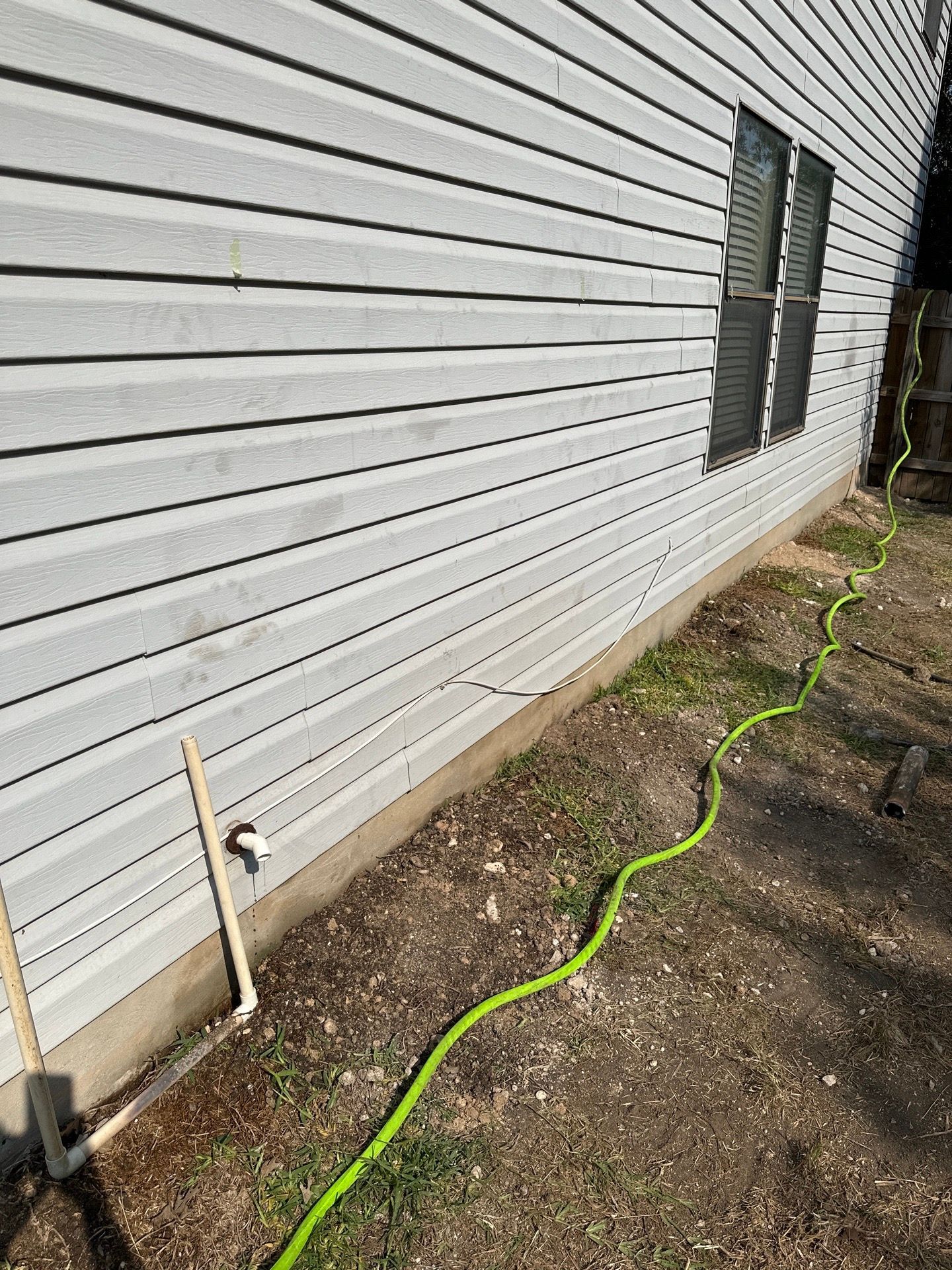 A green garden hose runs along a light gray siding of a building near the ground.