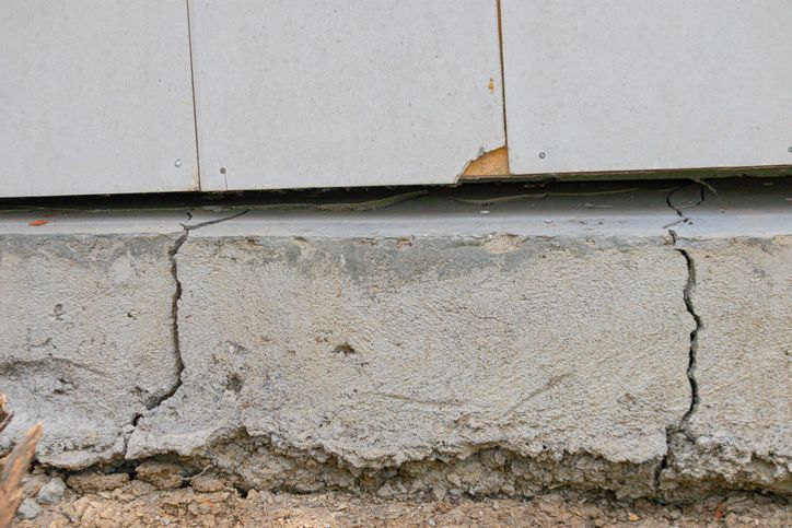 Close-up of a concrete foundation with vertical cracks. The foundation supports light-colored siding with a small piece missing. Close-up of a concrete foundation with vertical cracks. The foundation supports light-colored siding with a small piece missing.