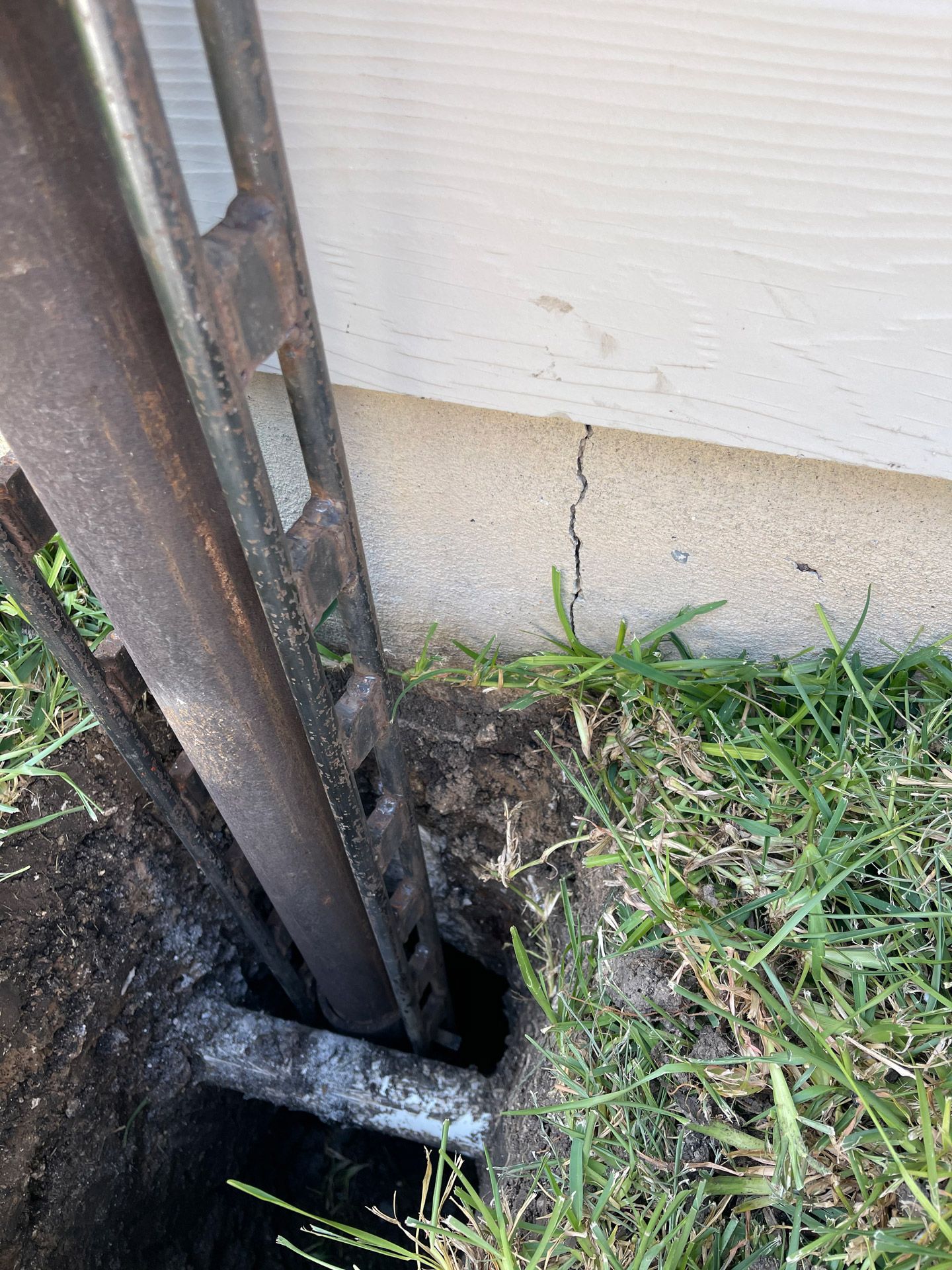 Damaged metal post set in a hole beside a house wall and grass, with cracked siding behind it