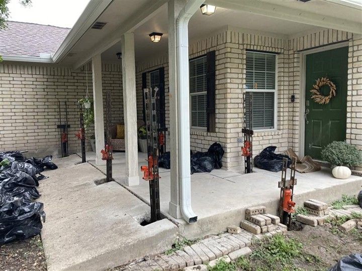 Exterior view of a porch with hydraulic jacks supporting the roof, concrete slab, and brick foundation; bags and bricks are nearby.