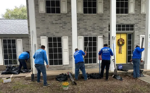 Five workers in blue shirts doing exterior work on a two-story house under construction
